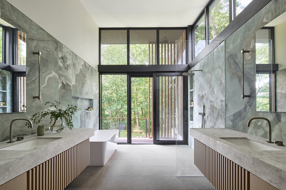 Modern bathroom with marble walls, dual sinks, a freestanding tub, large windows, and glass door that shows patio door opening to a balcony with greenery outside.
