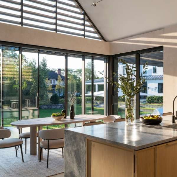Sunlit kitchen and dining area with modern patio doors, stylish black bar stools lined up at a marble island, light wood cabinetry, and large triangular windows and doors casting dramatic shadows, showcasing the seamless indoor-outdoor ambiance created by contemporary patio doors.