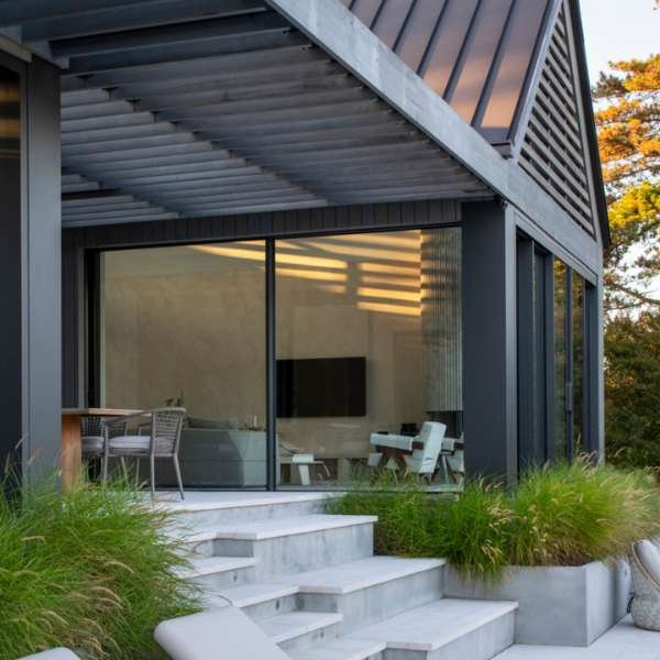 Modern home exterior featuring large sliding glass windows and doors, framed in sleek black metal, opening onto a minimalist patio with concrete steps, outdoor seating, and lush green landscaping—highlighting the seamless connection between indoor and outdoor spaces through contemporary windows and doors.