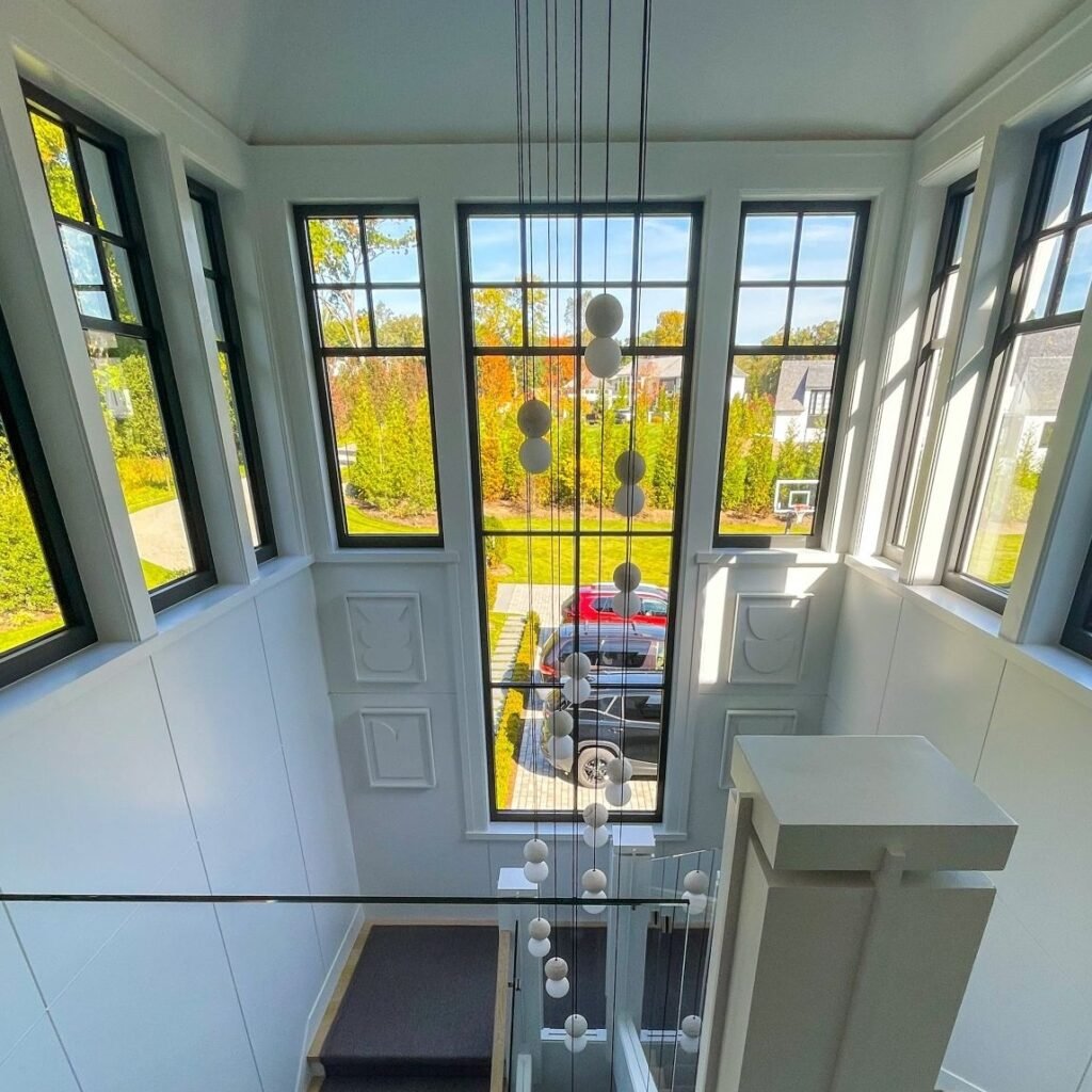 Interior view of a modern two-story foyer or stairwell with floor-to-ceiling black-framed windows offering views of a suburban neighborhood with autumn foliage. A dramatic multi-level pendant light fixture with spherical glass globes hangs from the high ceiling. The space features white walls with decorative molding panels, a glass and metal staircase railing, and built-in white cabinetry. Natural light floods the space, illuminating the clean, contemporary design