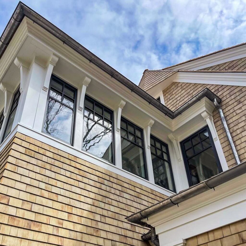 Exterior view of a modern house featuring a large black-framed window with grid pattern mullions flanked by two smaller matching windows. The facade combines white painted wood siding on the upper portion with natural cedar shake shingles below. Four white decorative columns support the overhanging roof structure, and the windows reflect the blue sky and clouds