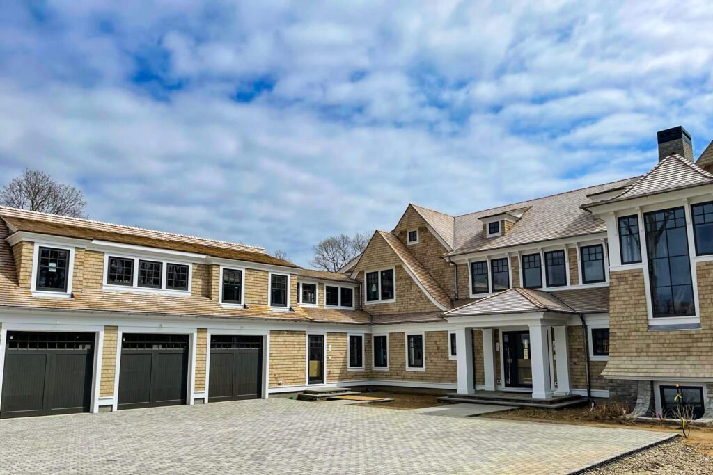 Exterior view of a modern big house featuring a large black-framed window with grid pattern mullions flanked by two smaller matching windows. The facade combines white painted wood siding on the upper portion with natural cedar shake shingles below. Four white decorative columns support the overhanging roof structure