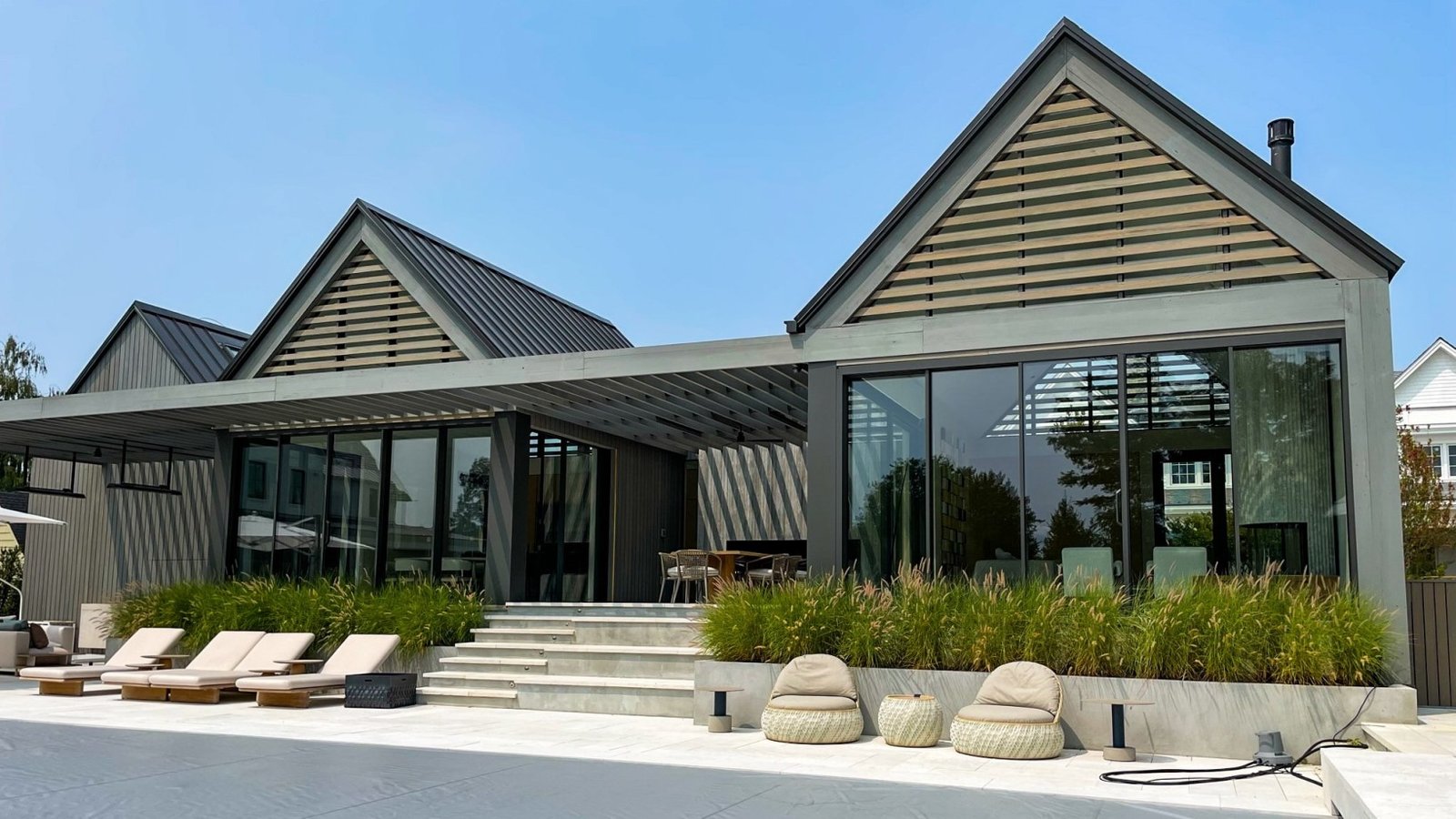 Modern home exterior featuring large sliding glass windows and doors, framed in sleek black metal, opening onto a minimalist patio with concrete steps, outdoor seating, and lush green landscaping—highlighting the seamless connection between indoor and outdoor spaces through contemporary windows and doors.