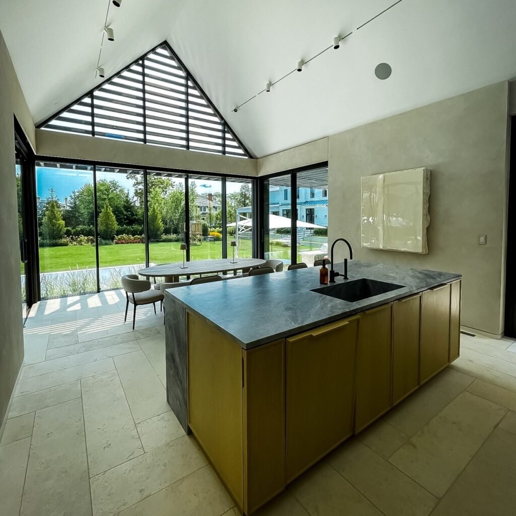 Modern home exterior featuring large sliding glass windows and doors, framed in sleek black metal, opening onto a minimalist patio with concrete steps, outdoor seating, and lush green landscaping—highlighting the seamless connection between indoor and outdoor spaces through contemporary windows and doors.