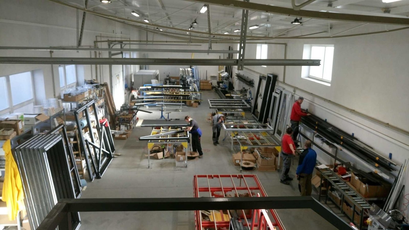 Three workers in red shirts assembling window frames at a manufacturing station.