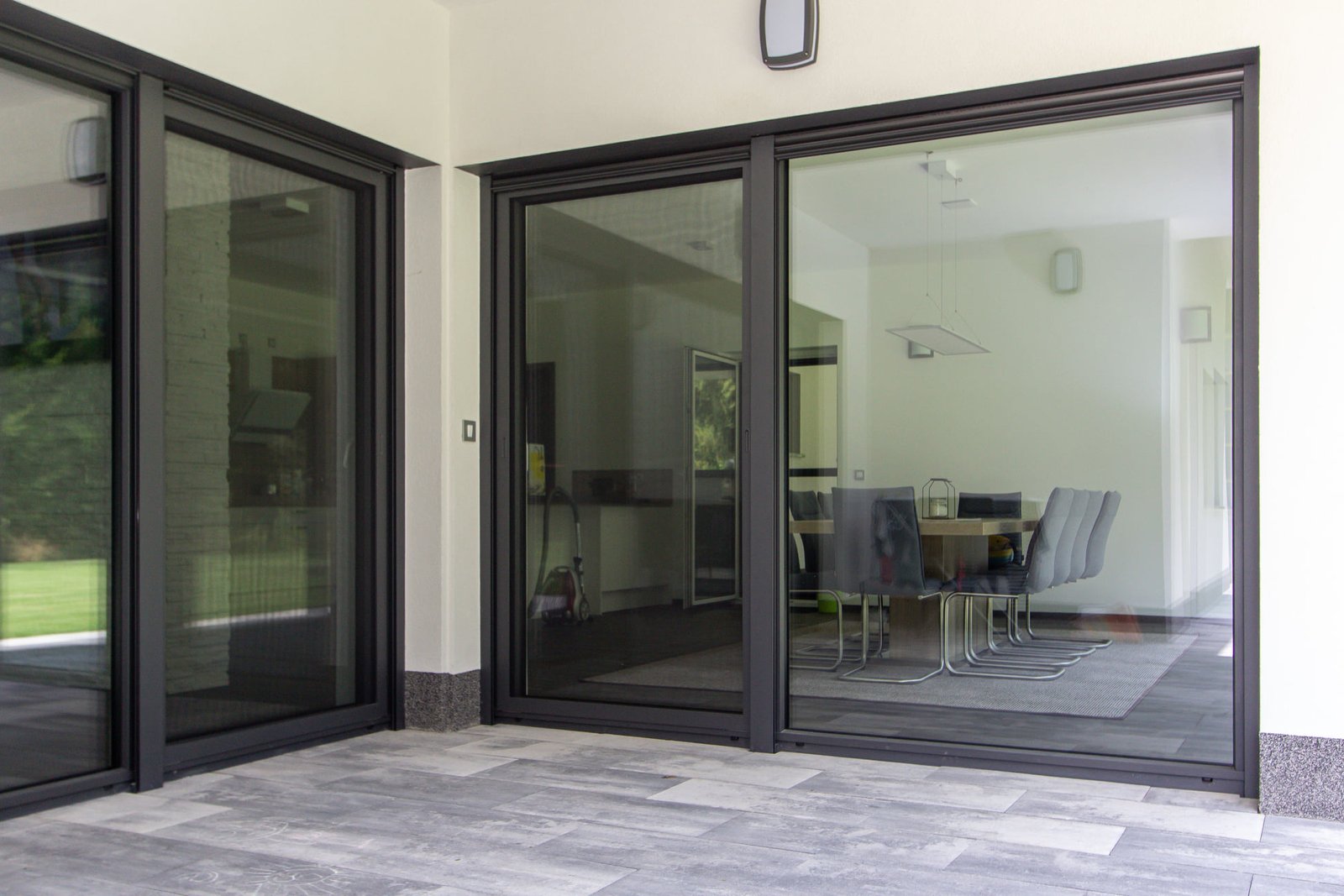 A modern European-style window with a sleek gray aluminum frame and double-glazed glass panels. One pane is slightly tilted open in a tilt-and-turn design, revealing a view of a sunny suburban landscape with green grass, trees, and houses in the distance. The clean interior and natural light emphasize the window’s energy-efficient and contemporary aesthetic.